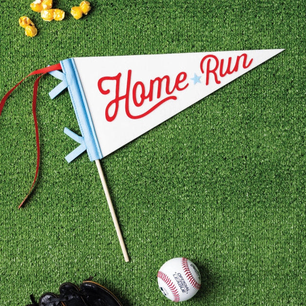 Felt “Home Run” baseball pennant on a wooden stick lying on bright green turf beside popcorn, baseballs, and a glove.