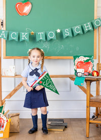 Young girl in school uniform holding felt “First Day of School” pennant in front of green chalkboard