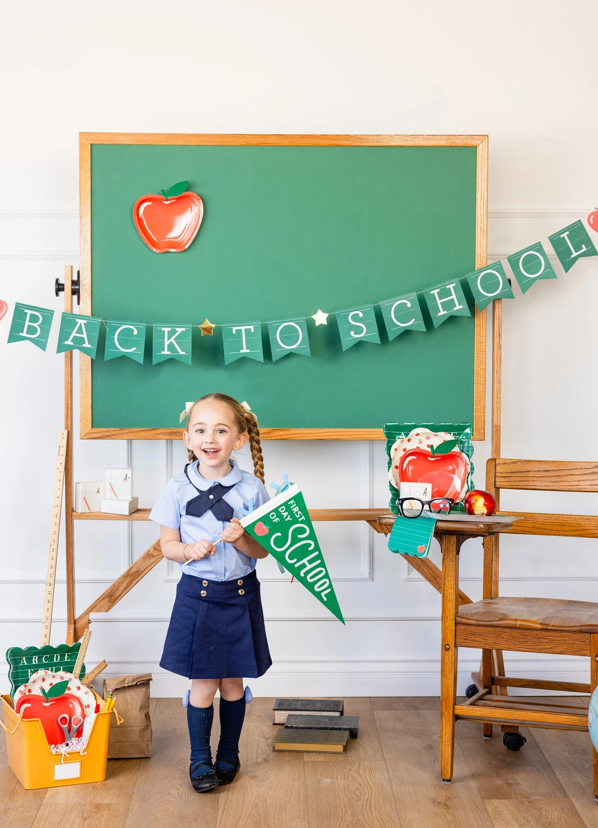 Girl smiling with green felt “First Day of School” pennant, surrounded by school-themed props