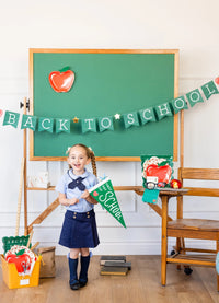 Girl smiling with green felt “First Day of School” pennant, surrounded by school-themed props