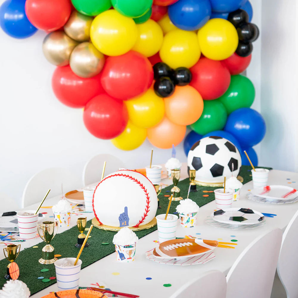 A festive kids' party table decorated with colorful balloons and sports-themed items including baseball and soccer cakes, mini trophies, and baseball party decorations.