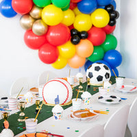 A festive kids' party table decorated with colorful balloons and sports-themed items including baseball and soccer cakes, mini trophies, and baseball party decorations.