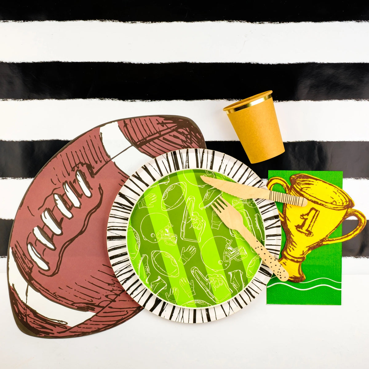 Football paper plate set up with a black and white striped table runner, golden trophy napkin, and wooden cutlery, ideal decorations for a tailgate party.