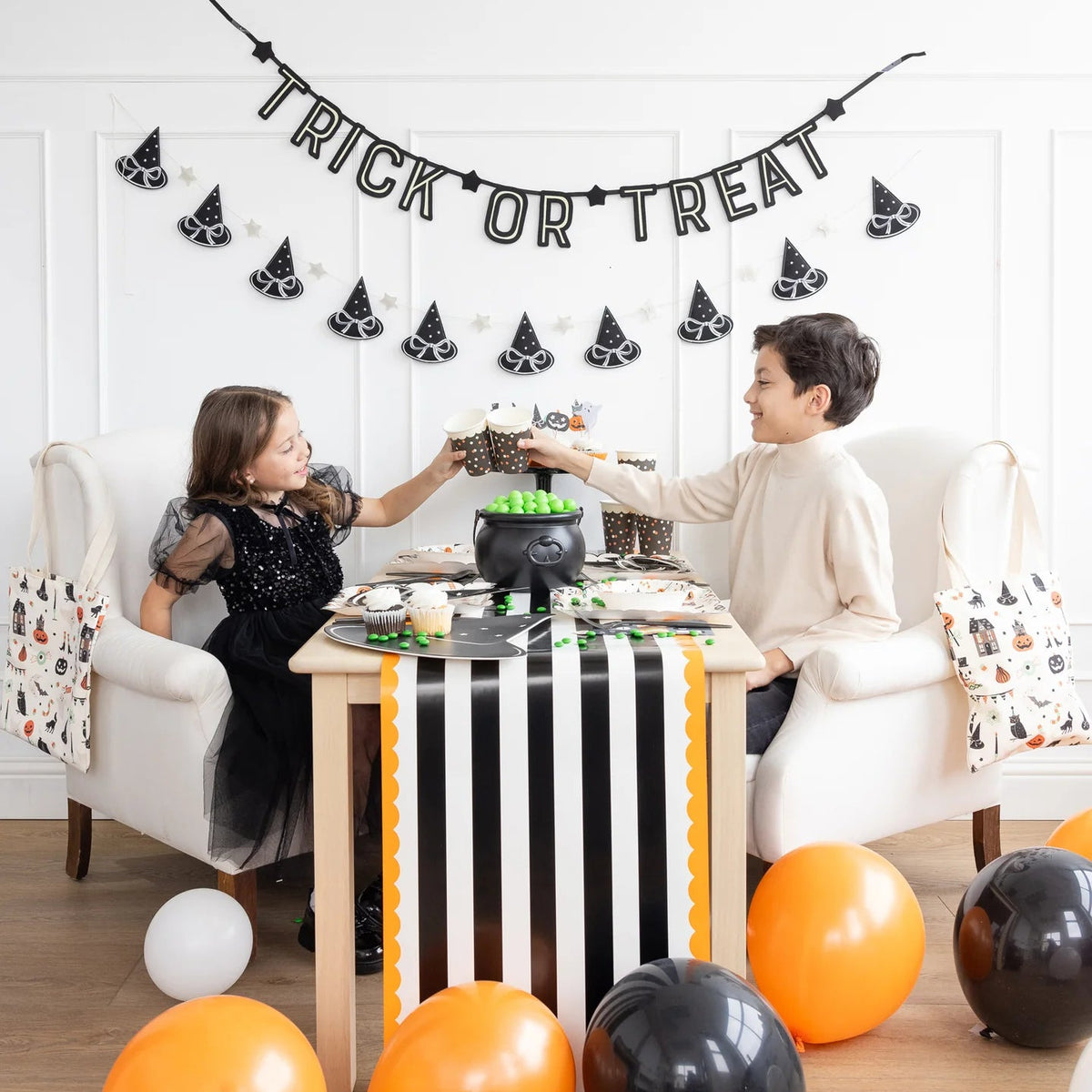 Two kids clink polka-dot Halloween cups over a table decorated with balloons and a cauldron candy bowl.