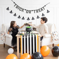 Two kids clink polka-dot Halloween cups over a table decorated with balloons and a cauldron candy bowl.