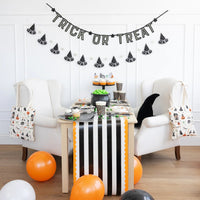 Halloween snack table with black-and-orange cups, striped runner, and Trick or Treat banner.