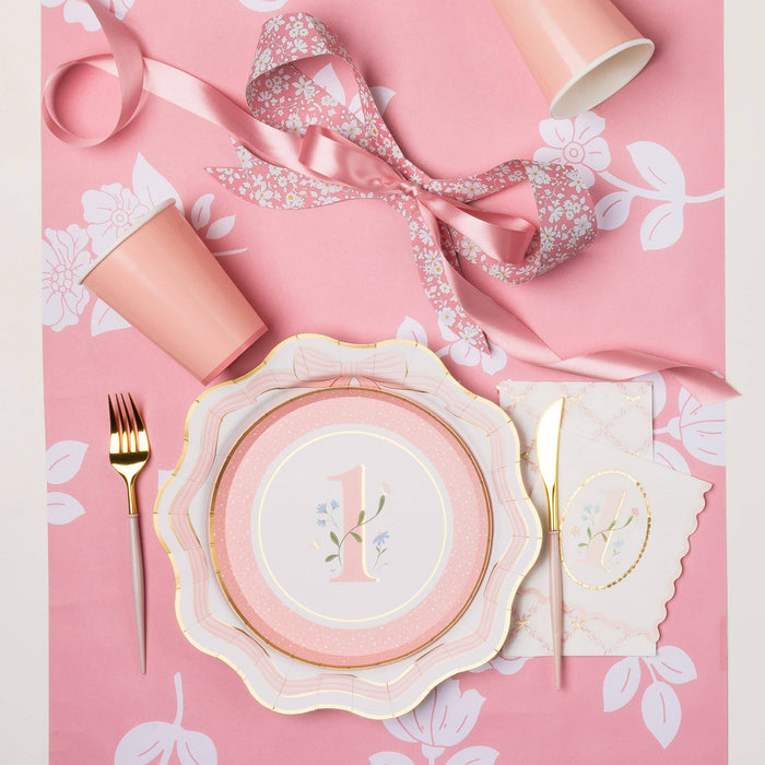 Overhead lifestyle shot of pink floral first birthday girl party table with floral number one plate, scalloped dinner plate, pink cup, gold cutlery and matching napkin on pink floral tablecloth with satin ribbon