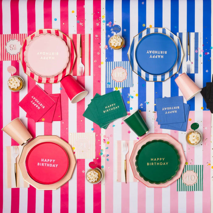 Overhead lifestyle shot of colorful birthday party table with assorted Happy Birthday plates, napkins and cups in pink, blue, and green on striped tablecloths