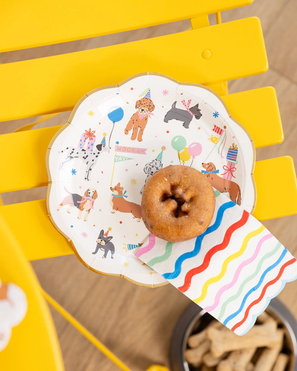 Dessert plate on a yellow chair with a maple donut and a folded rainbow napkin draped over the rim; dog-party pattern peeks from beneath.