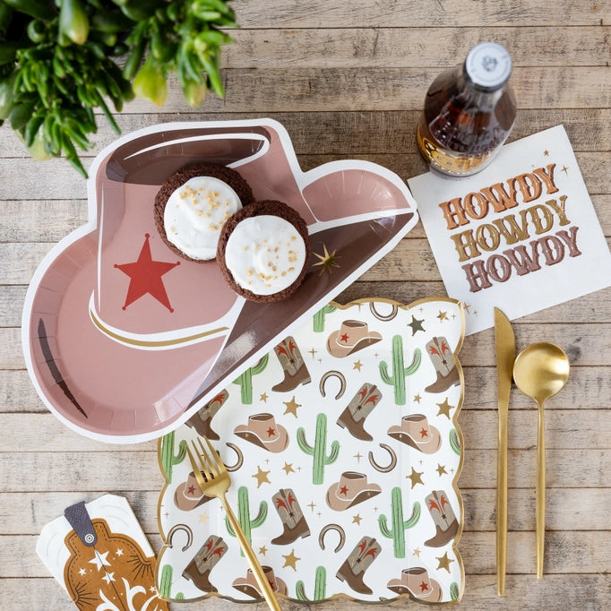Rustic wood table with cowboy hat plate, western icon plate, gold flatware, and HOWDY napkin beneath a soda bottle.