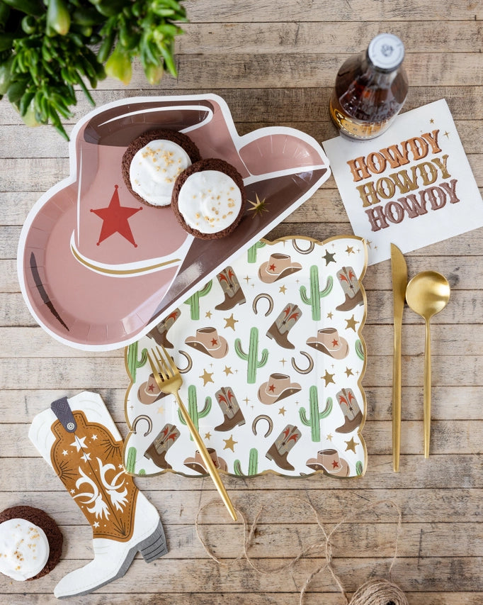 Rustic wood table with cowboy hat plate, square icon plate, “Howdy” napkin, gold flatware, twine, and cupcake.