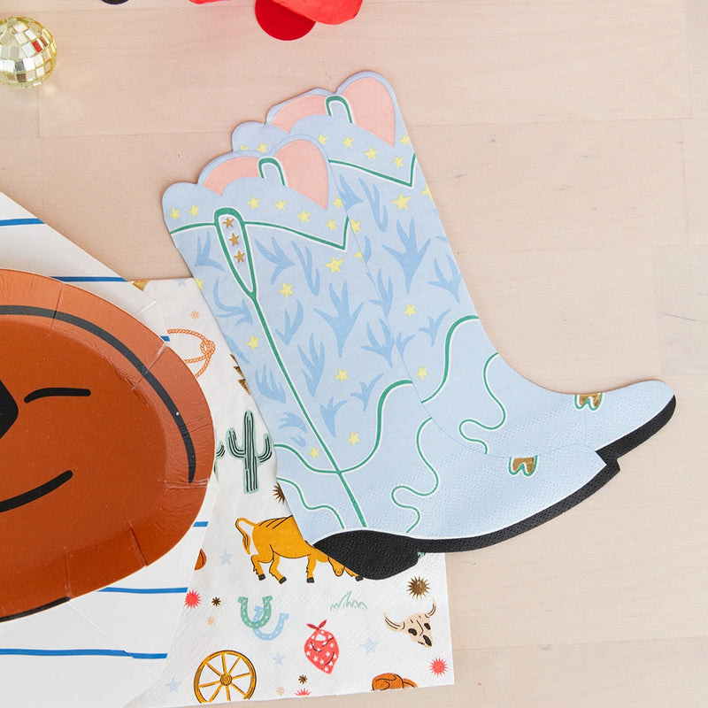 Detail – Boot Napkin Layer: Dusty‑blue boot napkins stacked on top of a cowboy‑motif napkin featuring cacti, bulls, and the word “HOWDY.”