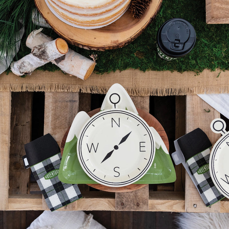 Overhead shot of a rustic wooden party table featuring a compass plate stacked over mountain-shaped plates, flanked by thermos napkins and cozy party details.