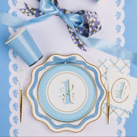 Overhead lifestyle shot of sky blue first birthday boy party table with floral number one cake plate stacked on bow dinner plate, matching napkin, blue cup, gold cutlery and blue satin ribbon on white scalloped placemat