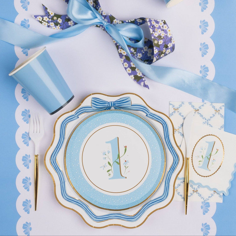 Overhead lifestyle shot of sky blue first birthday boy party table with floral number 1 plate, bow dinner plate, blue cup, gold cutlery and blue ribbon on white scalloped placemat