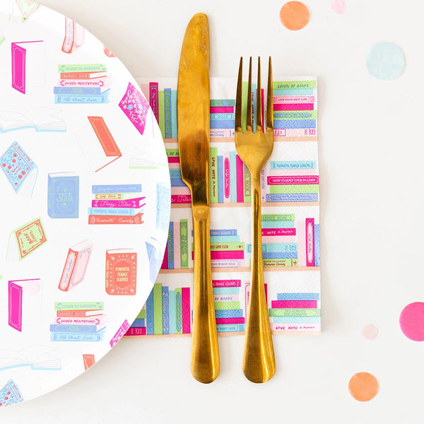Gold knife and fork resting on folded bookshelf paper napkin beside pastel book-pattern dinner plate.
