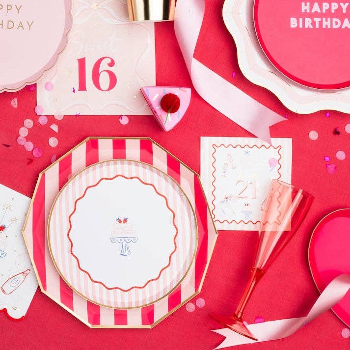 Overhead lifestyle flat lay of pink 21st birthday party table with striped birthday cake plates, Happy Birthday plate, Cheers to 21 cocktail napkins, gold cup and confetti on red tablecloth