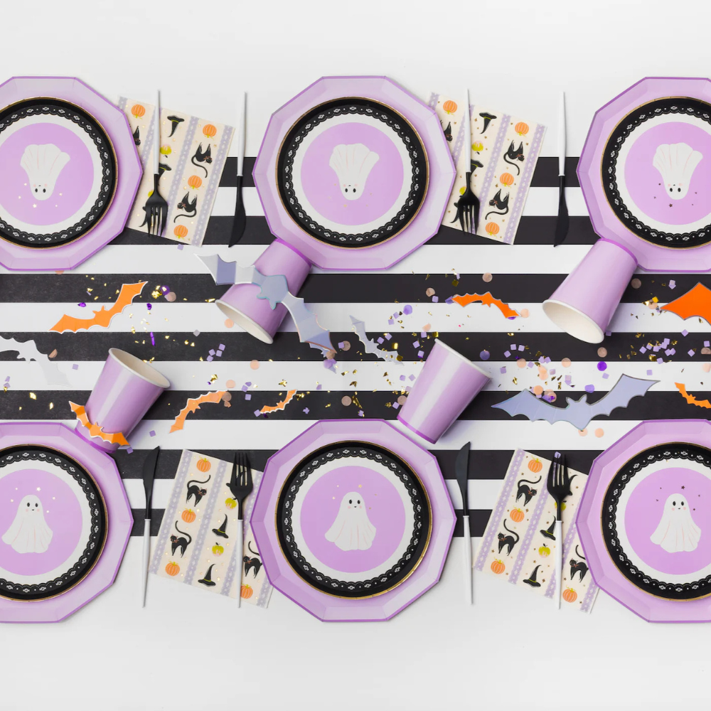 Styled Halloween table with ghost plates, purple cups, and Frills & Thrills Halloween napkins featuring pumpkins, cats, and witch hats.