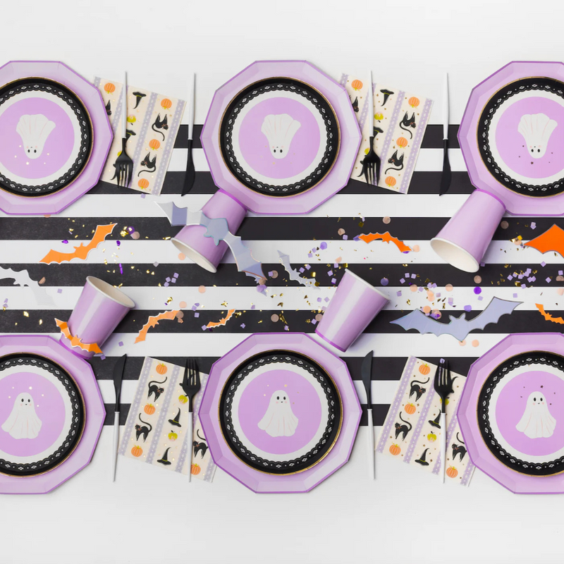 Styled Halloween table with ghost plates, purple cups, and Frills & Thrills Halloween napkins featuring pumpkins, cats, and witch hats.