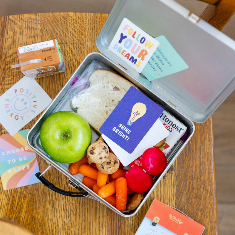 Open lunchbox on a table with colorful cards and a “Shine Bright” note inside.