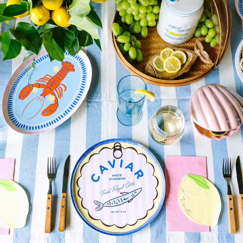 A close-up of the Le Dinner Caviar Can Dinner Plate styled with grapes and glassware on a striped tablecloth.
