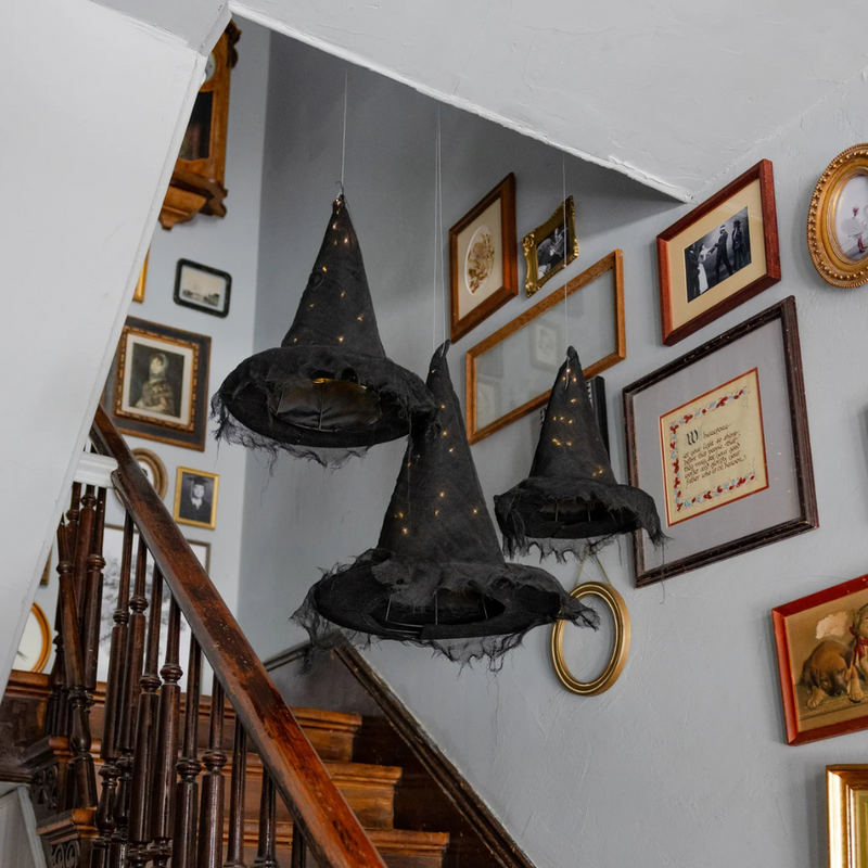 Close-up of three black light-up witch hats suspended over a staircase with framed pictures.