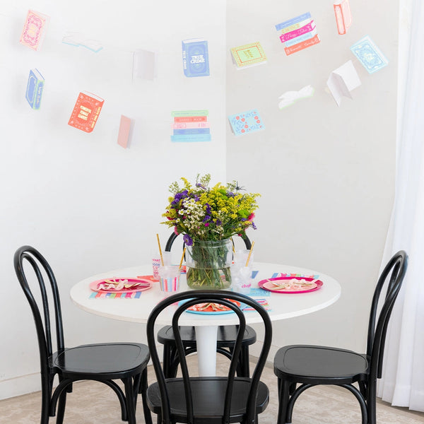 Round table set for four under pastel book garland; bright plates and floral centerpiece create a cozy book club scene.