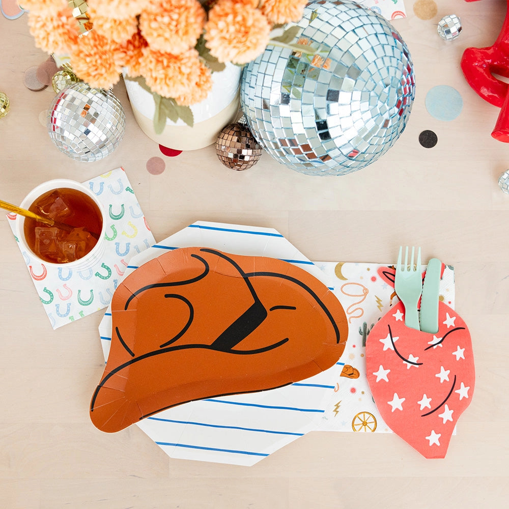 Close‑up of a single cowboy‑hat plate resting on a blue‑striped octagon plate; a cup of iced tea and rainbow horseshoe napkin sit beside silver and rose‑gold disco balls.