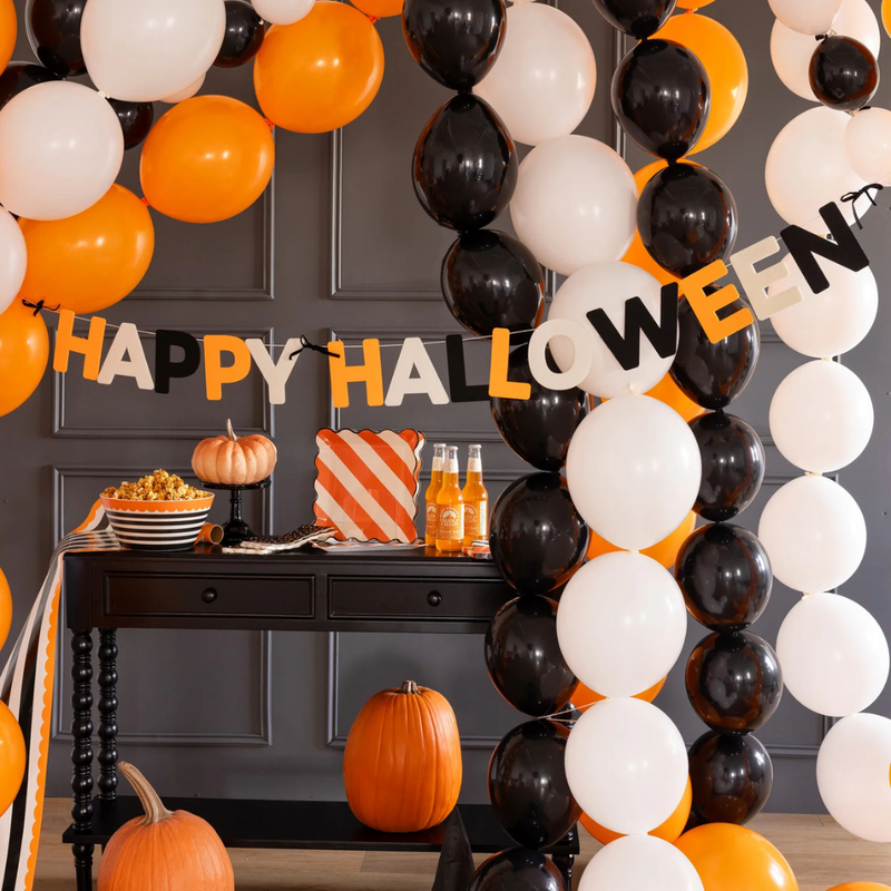 Felt & Bows Happy Halloween Banner hanging across a black wall with orange, white, and black balloons for decoration.