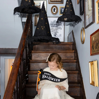 Child holding a Happy Halloween sign beneath illuminated hanging witch hats.