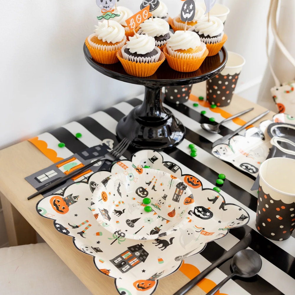Close-up of cupcakes and Halloween tableware on black and white striped paper table runner with orange scalloped edges.