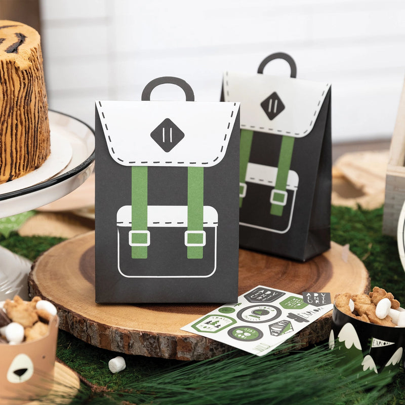 A rustic party table setup featuring black and green paper backpack favor bags, mini bear snack cups, and forest-themed decor.
