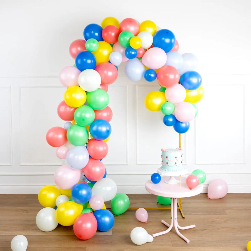 Floor‑to‑ceiling rainbow balloon garland framing a white wall, pastel cake on pink pedestal table in foreground.