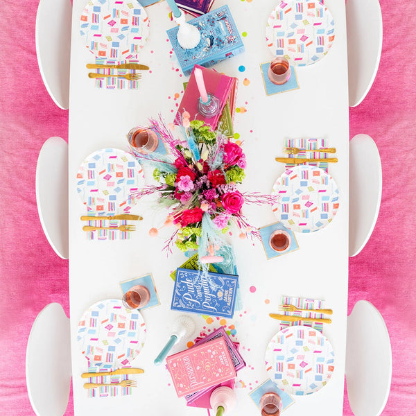 Overhead view of bright pink-accent book club table with book-pattern dinner plates, novels, floral centerpiece, and pink drink cups