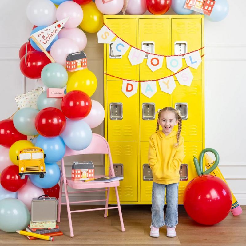 Little girl smiling in front of yellow lockers and colorful balloon garland with school bus party decorations
