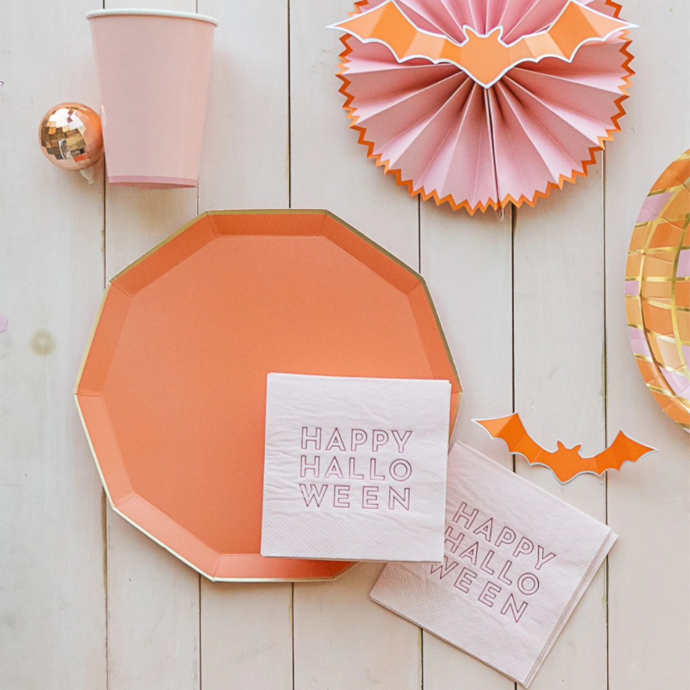 Close-up of pink cocktail napkin with “Happy Halloween” text in bold lettering beside an orange plate and cup.