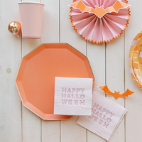Close-up of pink cocktail napkin with “Happy Halloween” text in bold lettering beside an orange plate and cup.