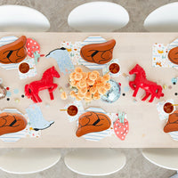 Overhead view of a long wooden dining table set for a kids’ western‑themed party—rust‑orange cowboy‑hat plates atop striped chargers, red toy horses as centerpieces, disco‑ball ornaments, striped cups with yellow straws, and patterned napkins scattered around.