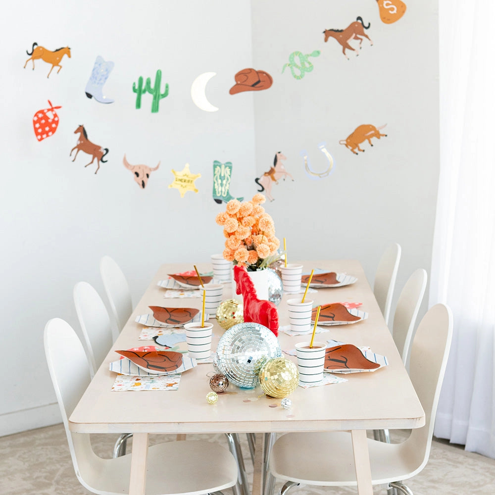 Long dining table set with rust‑orange cowboy‑hat plates, striped cups and disco‑ball centerpieces under a western‑themed garland.