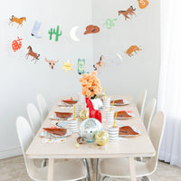 Long dining table set with rust‑orange cowboy‑hat plates, striped cups and disco‑ball centerpieces under a western‑themed garland.