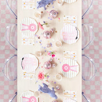 Overhead view of a white table set with octagon plates, pink show ribbon napkins, gingham runner, disco balls, and horse-print napkins for a cowgirl party.