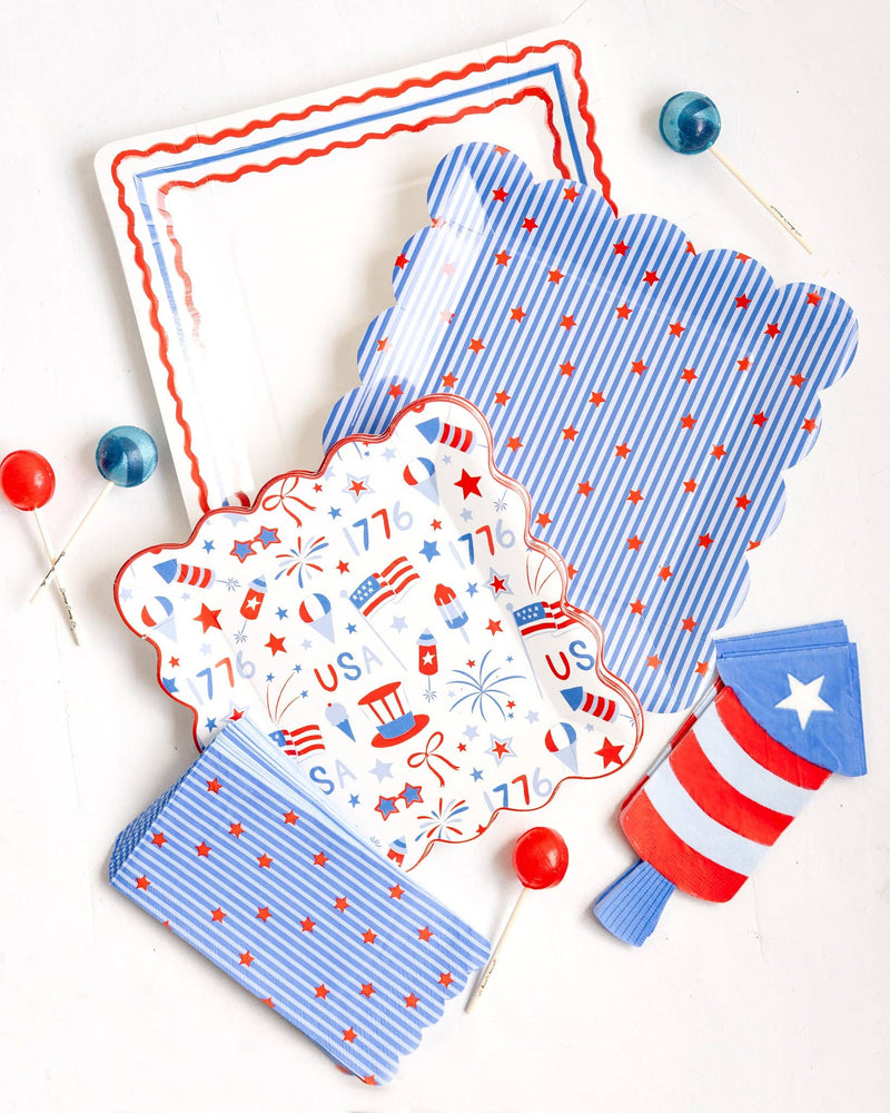 Flat lay of a patriotic party collection including a cream square border dinner plate, blue striped scalloped plate, 4th of July icons scalloped plate, firecracker shaped die cut napkins, blue star napkins, and red and blue lollipops on a white background