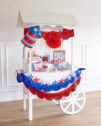 White party cart decorated for 4th of July with layered blue and red metallic fringe banners, 3D star garland, firecracker shaped balloon, paper fans, and patriotic party supplies