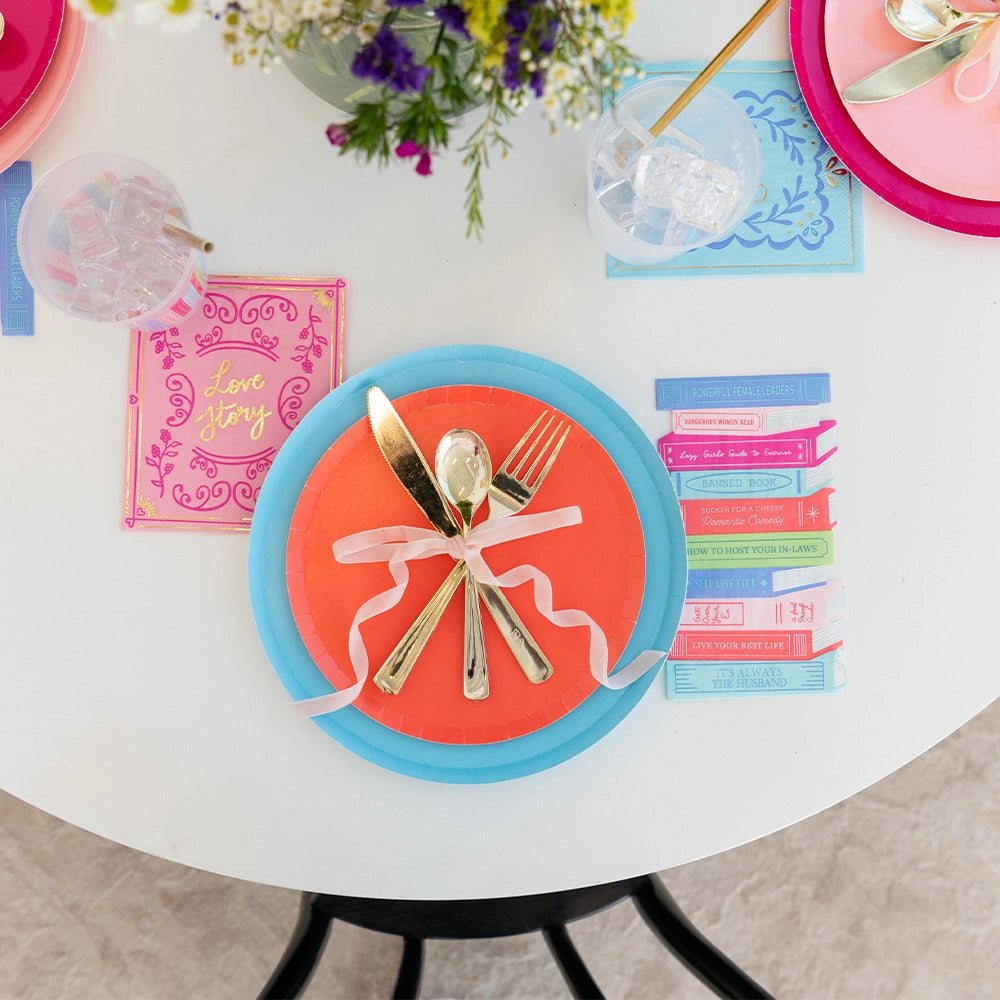 Orange and teal plate place setting with gold utensils tied by ribbon; pastel book napkin beside iced drink.