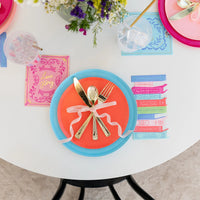 Orange and teal plate place setting with gold utensils tied by ribbon; pastel book napkin beside iced drink.