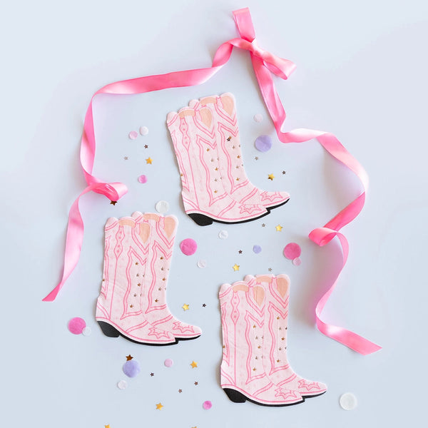 Three pink boot-shaped napkins with gold star accents, arranged on a light-blue backdrop with pink ribbon curls and pastel confetti—perfect for a cowgirl-themed celebration.