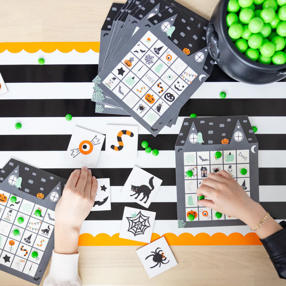 Hands playing Halloween-themed bingo on a black and white striped paper table runner with orange scalloped edges.
