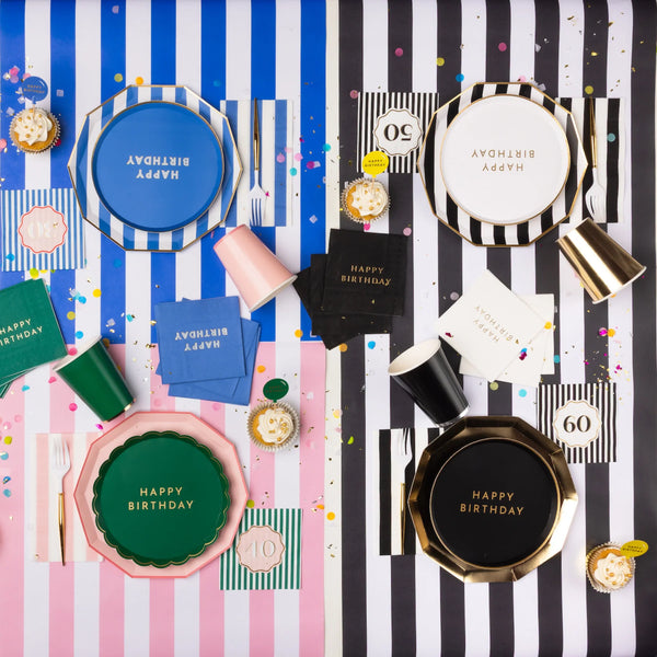 Overhead lifestyle shot of elegant black and white 60th birthday party table with black Happy Birthday plates, gold cups, striped napkins and confetti alongside colorful milestone birthday setups