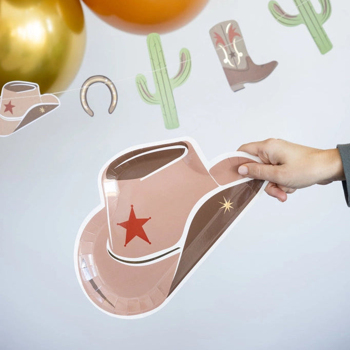 Hand holding cowboy hat plate in front of western garland with cacti, boots, horseshoes, and brown‑gold balloons.