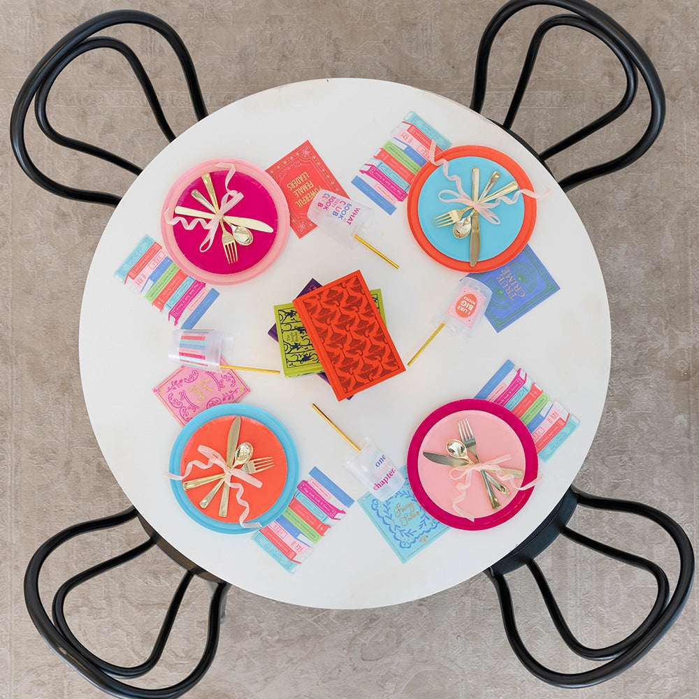 Overhead view of four-place book club table—bright plates, gold cutlery, and stacked book guest napkins.